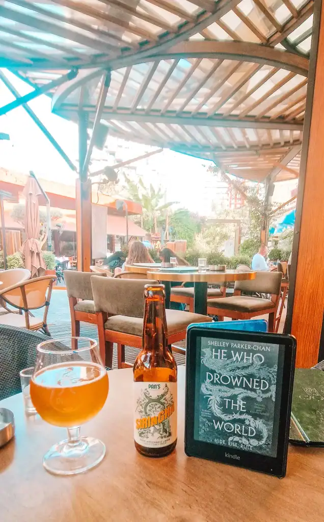 A Kindle e-reader displaying the cover of He Who Drowned the World by Shelley Parker-Chan sits next to a bottle of craft beer at an outdoor café; a cozy scene for reading books about China.