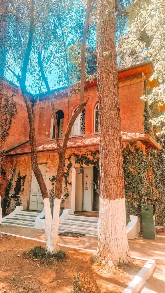 A vine-covered building with rustic reddish walls and white steps, shaded by tall trees—home to one of Tirana’s most intriguing museums.