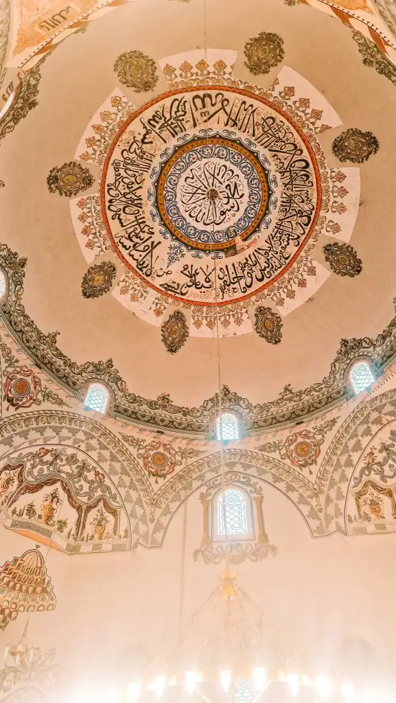 Ornate ceiling of a mosque featuring detailed Islamic calligraphy and floral patterns in earthy and golden tones, with light filtering through small arched windows.
