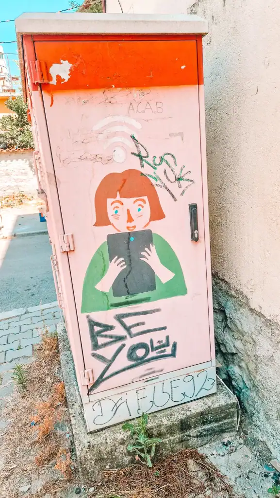 A painted utility box shows a cartoon girl holding a tablet, with graffiti around her and the words “Be You” in black lettering—Tirana’s quirky urban art popping up where you least expect it.