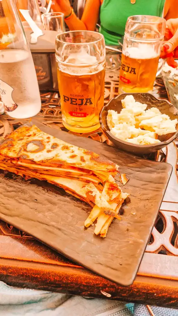 Close-up of a traditional meal featuring layered flatbread, a bowl of white cheese, and mugs of Peja pilsner beer on a patterned metal table during a relaxed stop on a Kosovo day trip.
