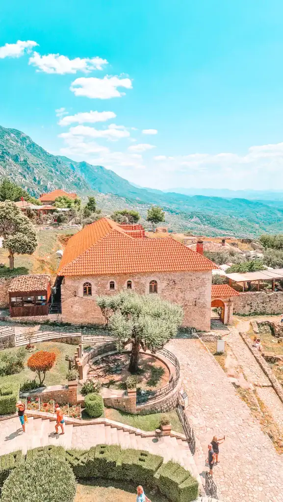 A scenic view of Krujë Castle with stone pathways, manicured gardens, and red-tiled roofs surrounded by green hills and mountains. Tourists walk up the stone stairs and take selfies under a bright blue sky dotted with fluffy clouds.