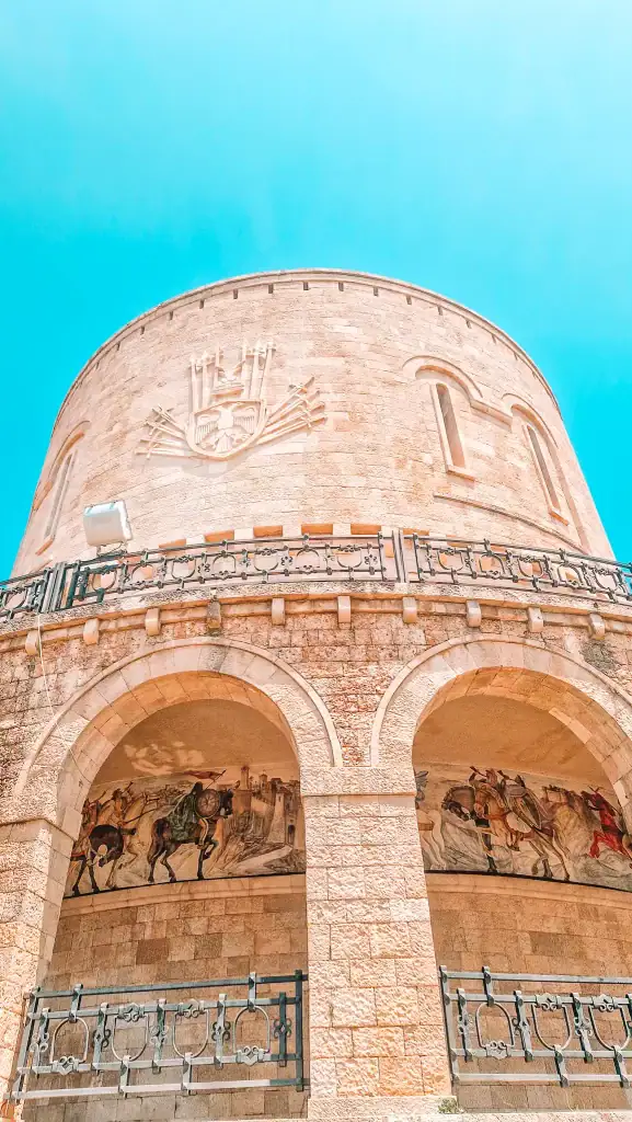 Upward view of the Skanderbeg Museum in Krujë, a round stone structure featuring engraved Albanian crest and a fresco of medieval warriors under grand arches.
