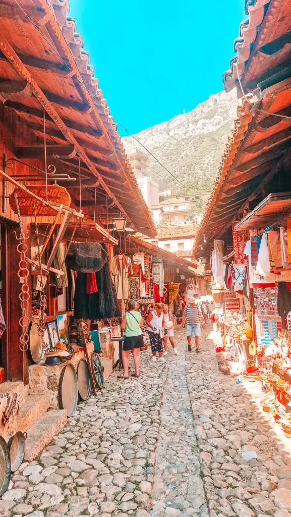 Narrow cobblestone street lined with wooden market stalls in Krujë, selling traditional Albanian crafts, embroidered garments, musical instruments, and souvenirs.