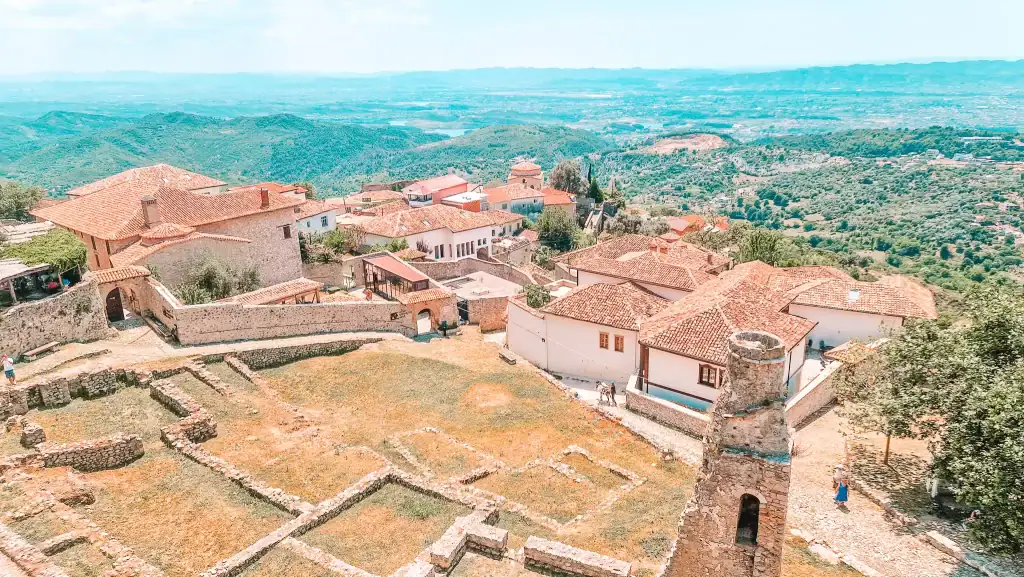 Scenic hilltop view over the red-tiled roofs and ruins of Krujë Castle, with mountains and valleys stretching out beneath a bright sky.