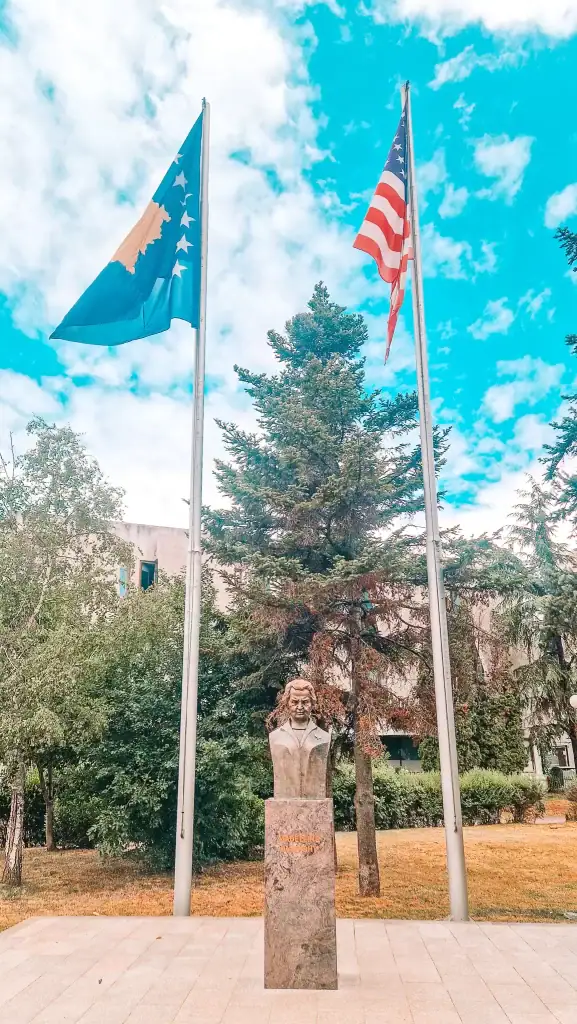 A bronze bust of Madeleine Albright stands between the Kosovo and American flags in a public square lined with trees and greenery under a vivid blue sky.