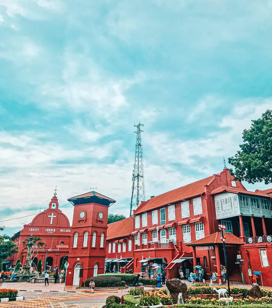 The iconic red buildings of Malacca’s Dutch Square, including Christ Church and the Stadthuys, stand out against a blue sky with tourists milling about the colorful plaza.