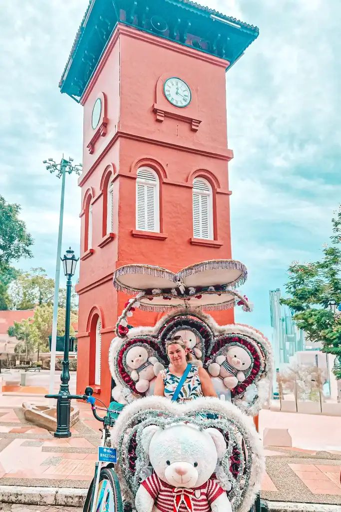Em smiles from a vibrantly decorated trishaw in Malacca, adorned with oversized teddy bears and heart-shaped designs, parked in front of the historic red clock tower.