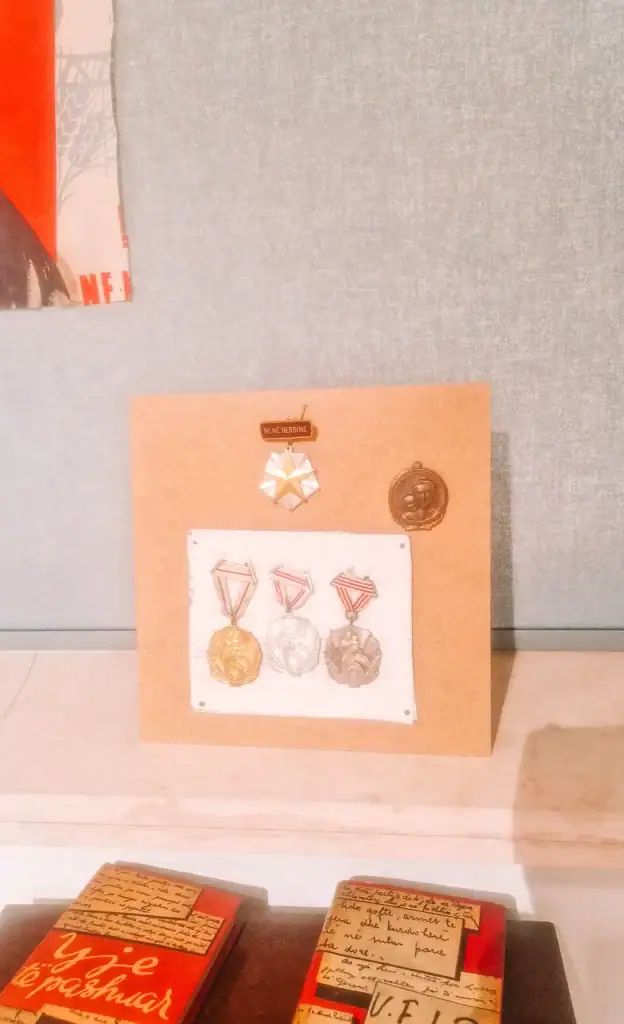 : A board of military-style medals and honor pins labeled “Nënë Heroinë,” showcasing national recognition for women’s contributions in Albanian history.
