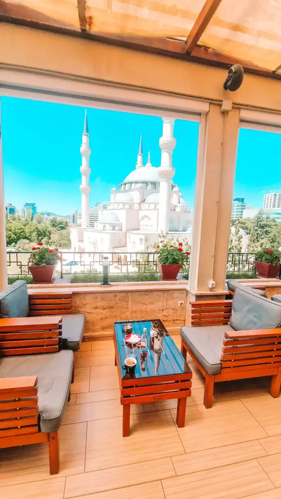 Rooftop seating at Monarc Café in Tirana with plush wooden lounge chairs and a stunning panoramic view of the Grand Mosque. Red geraniums line the railing, making this one of the best cafes in Tirana for a scenic coffee break.