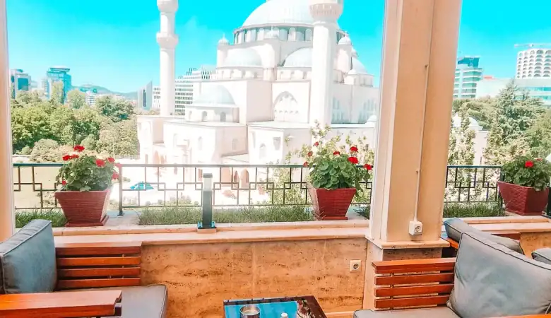 Rooftop seating at Monarc Café in Tirana with plush wooden lounge chairs and a stunning panoramic view of the Grand Mosque. Red geraniums line the railing, making this one of the best cafes in Tirana for a scenic coffee break.