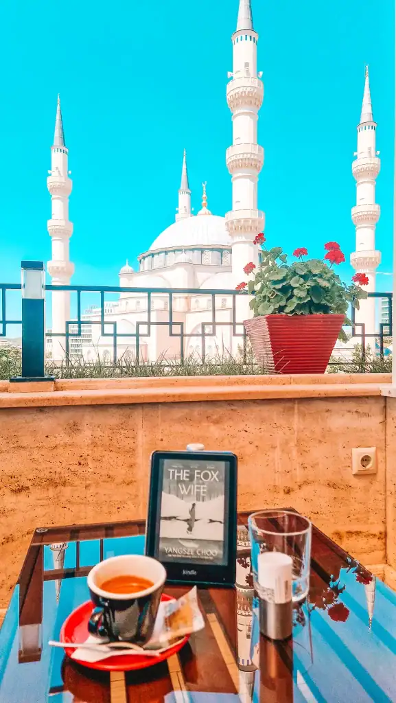 Espresso and Kindle on a sleek glass table at Monarc Café in Tirana, with “The Fox Wife” by Yangsze Choo open against the striking backdrop of the Grand Mosque's minarets. A peaceful, picturesque spot for reading and sipping in the heart of Tirana.