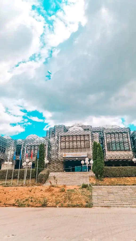 The National Library of Kosovo in Pristina features a striking modernist design with a metal lattice facade and domed roofs, standing under a partly cloudy sky with flags waving out front.