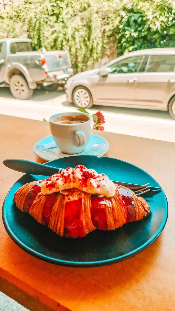 Close-up of a vibrant red croissant topped with whipped cream and paired with an espresso, sitting by a window overlooking parked cars. This treat is a standout at Nefeli, a sweet stop among the best cafes in Tirana.