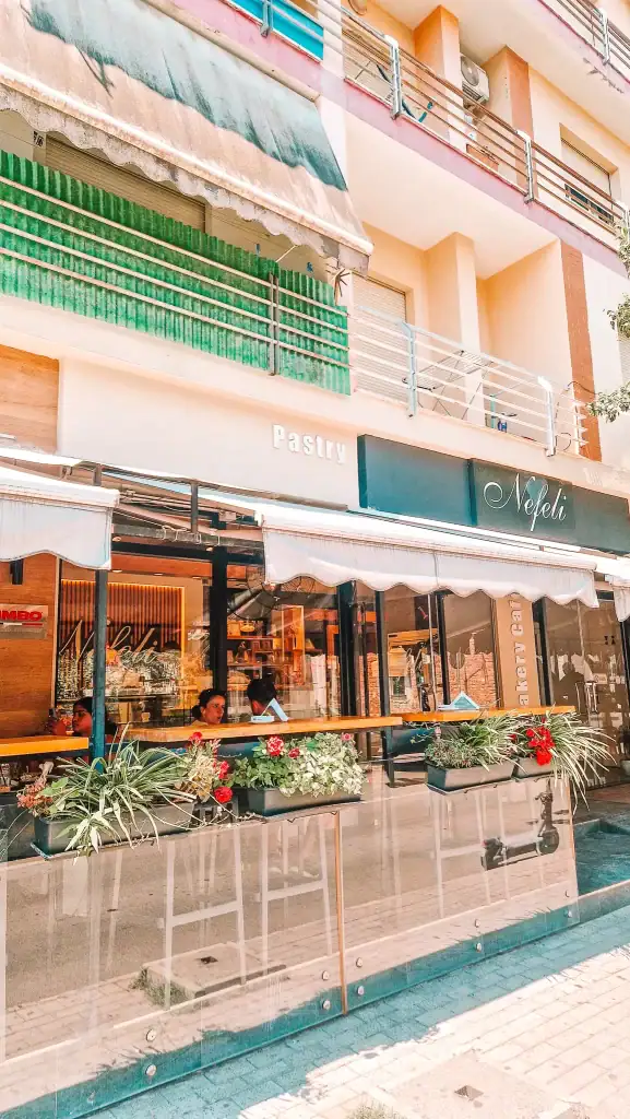 Exterior view of Nefeli Pastry in Tirana, with flower boxes, glass railing, and customers seated at the window bar. Its modern look and delicious menu have made it a popular name among cafes in Tirana.