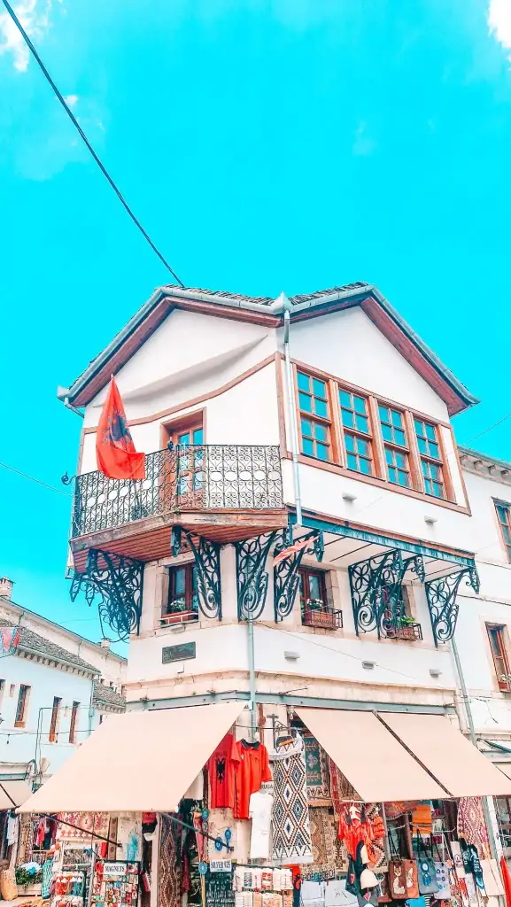 White Ottoman-style building with decorative balconies and an Albanian flag, sitting above a street stall packed with local souvenirs, rugs, and clothing.