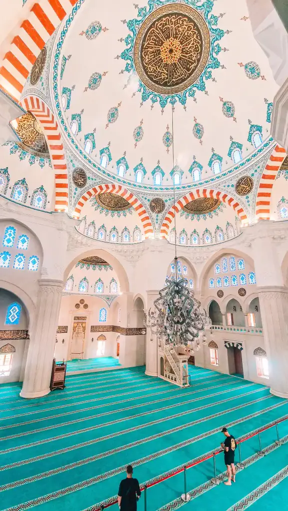 The stunning interior of the mosque featuring turquoise carpet, soaring white arches, stained-glass windows, and a richly decorated dome with Islamic-style calligraphy and patterns.