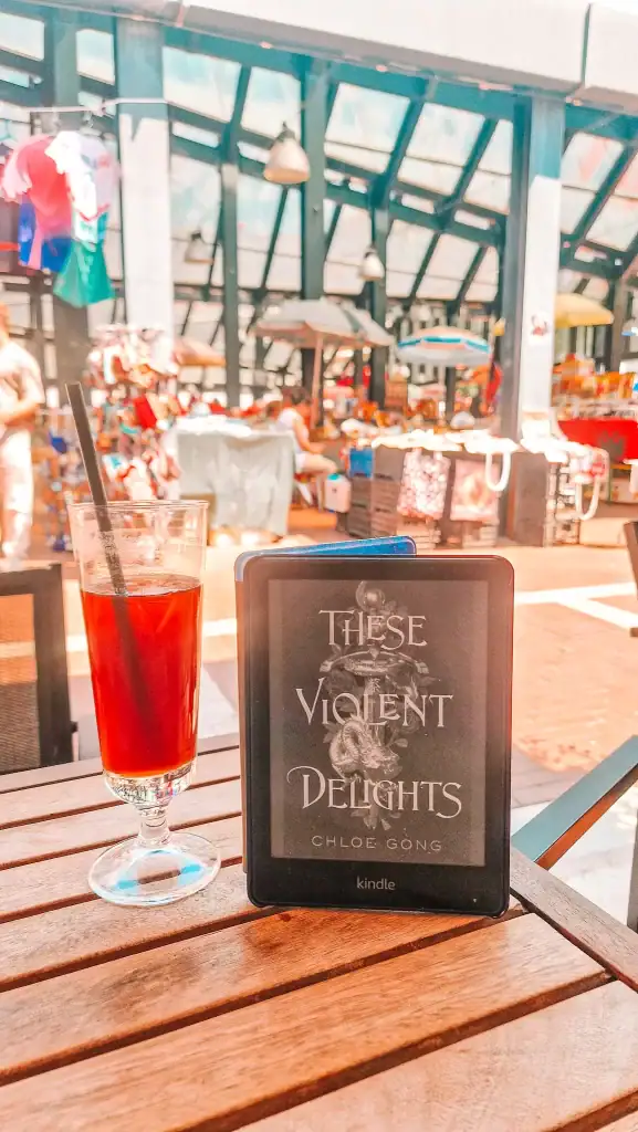 A Kindle showing These Violent Delights by Chloe Gong stands on a café table next to a cold drink, highlighting books about China in a lively market setting.