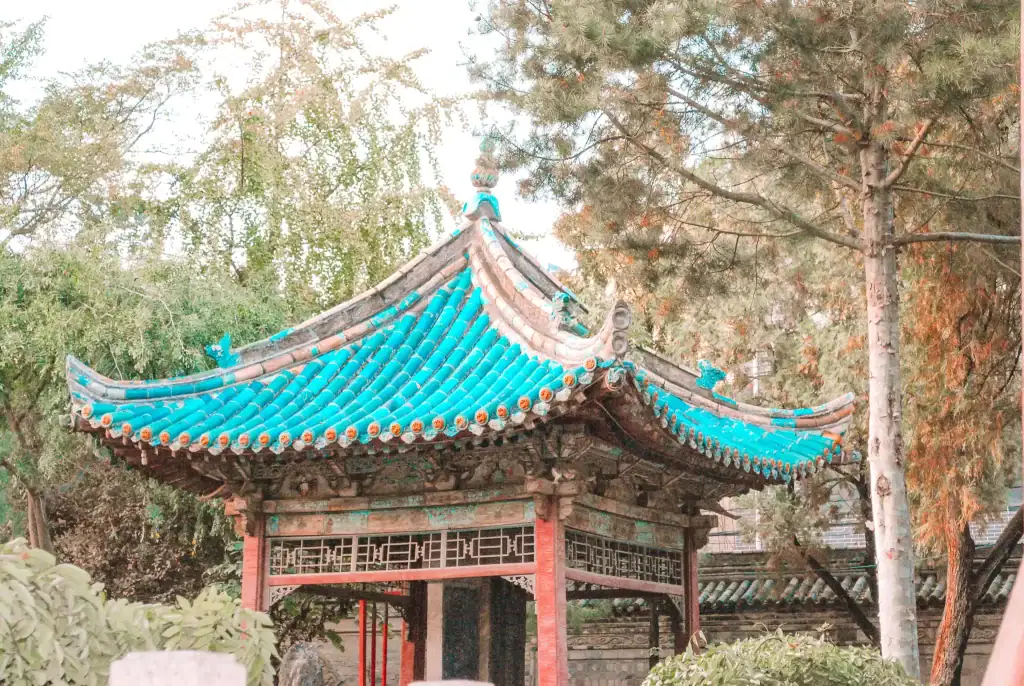 A traditional Chinese pavilion with turquoise roof tiles stands surrounded by lush trees, likely part of an ancient cultural site.