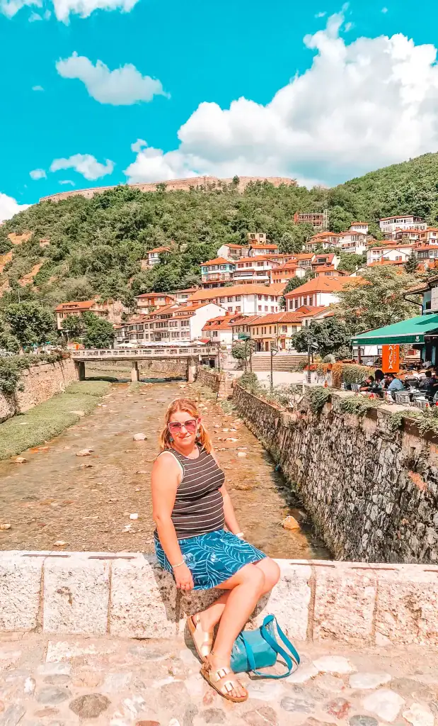 Em poses on a stone bridge in Prizren, Kosovo, with traditional red-roofed houses climbing a green hillside behind her. The shallow stone-filled river flows beneath the bridge as people dine at cafes along the riverside.