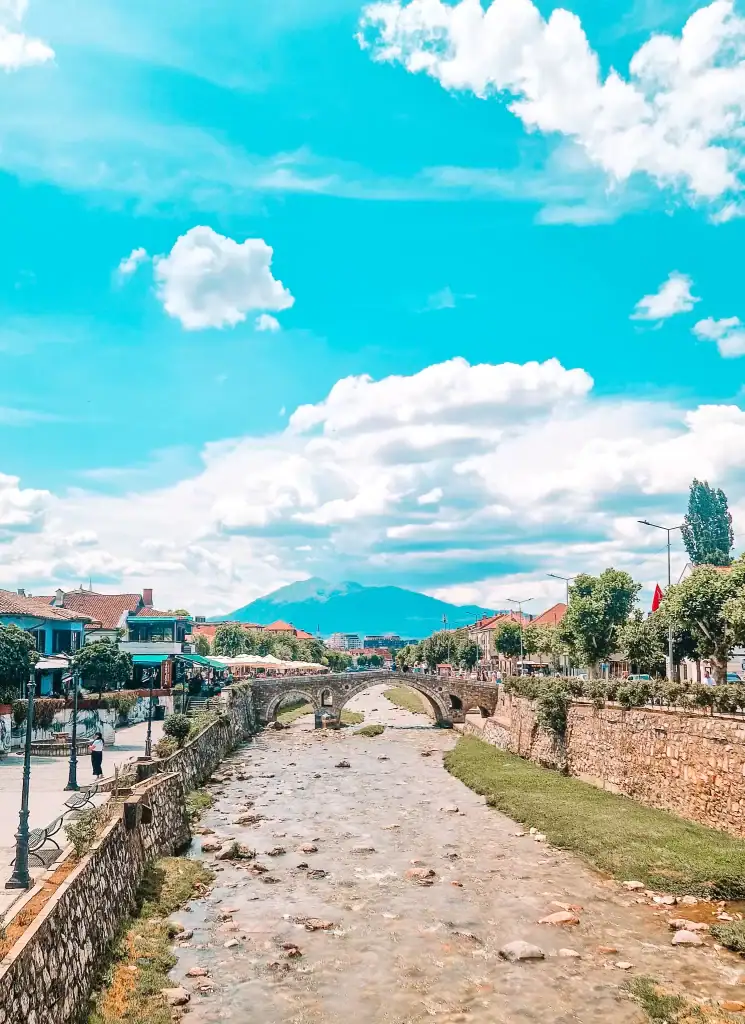 A scenic view of a stone bridge crossing a shallow, rocky river lined with cafés and buildings, with mountains visible in the background on a bright, clear day—ideal for a Kosovo day trip from Tirana.