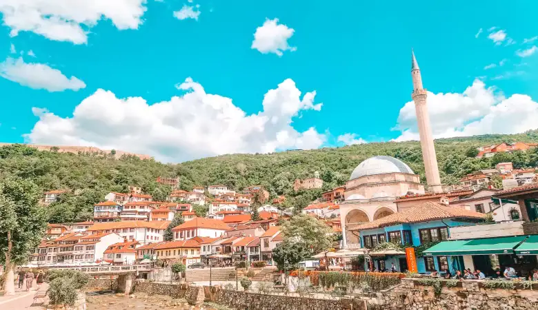View of the old town in Prizren, Kosovo, with a prominent white-domed mosque and tall minaret surrounded by red-roofed buildings nestled into a green hillside. This colorful and historic setting is a highlight of a Kosovo day trip from Tirana.