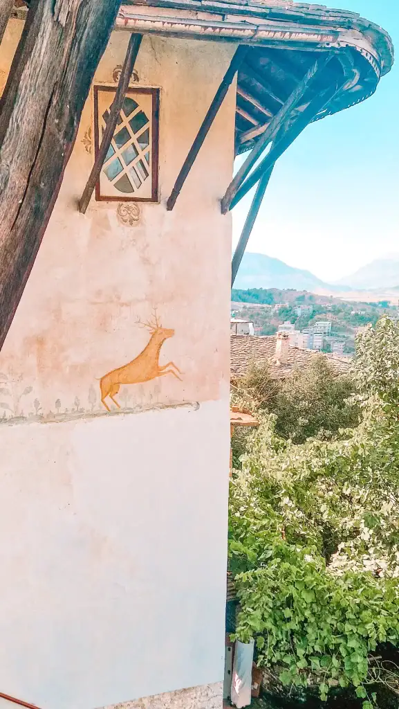 A close-up of a faded deer mural on the outer wall of Skenduli House, with a view of Gjirokaster’s rooftops and mountains beyond.