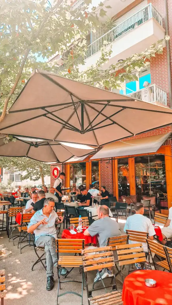 Bustling outdoor patio of Sophie Café in Tirana, with large umbrellas shading wooden tables draped in red tablecloths. Locals chat and sip coffee in the midday sun, capturing the social heart of one of the best cafes in Tirana.