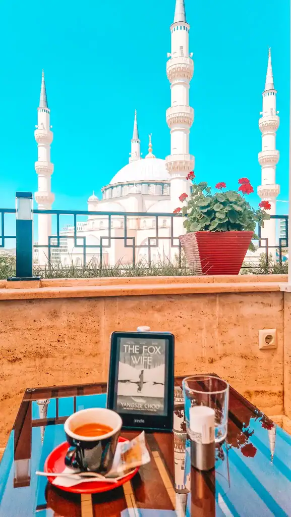 A Kindle displaying The Fox Wife by Yangsze Choo sits next to a cup of espresso on a table with a mosque in the background.