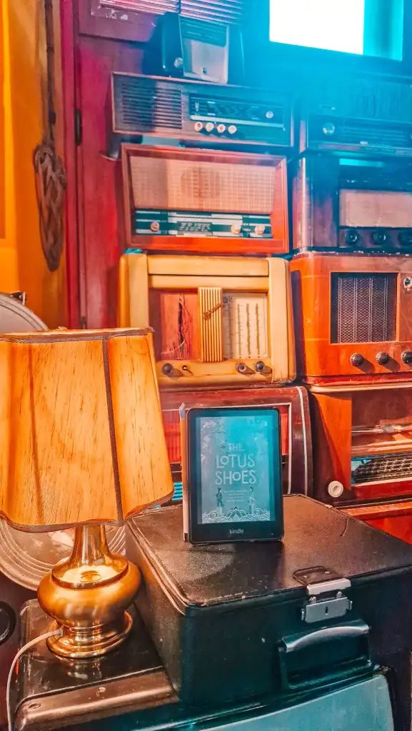 A cozy indoor scene with a Kindle showing The Lotus Shoes by Marah Ellis Ryan, resting beside a lamp and vintage radios.