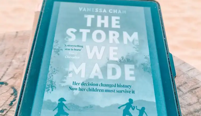 A Kindle on a beachside tree stump displays The Storm We Made by Vanessa Chan, with waves rolling in behind. This gripping debut is among the bold new books about Malaysia, focusing on a mother’s secret wartime choices during Japanese-occupied Malaya.