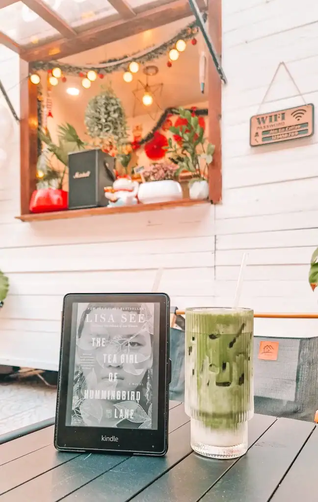 A Kindle displaying The Tea Girl of Hummingbird Lane by Lisa See stands next to an iced matcha latte in a café decorated with plants and lights