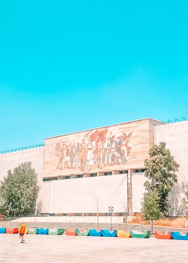 The large mosaic mural “The Albanians” on the National History Museum’s facade shows heroic figures from different eras holding weapons and flags, with rainbow-colored benches in the foreground—one of Tirana’s most iconic and symbolic landmarks.