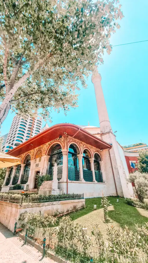 The Et’hem Bey Mosque peeks through leafy trees, showcasing its ornate arched porch, red-tiled roof, and tall minaret—an essential stop for anyone exploring Tirana in 2 days.