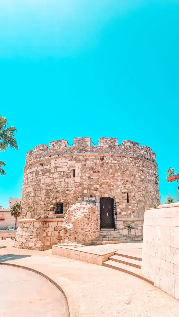 Circular stone tower of the Venetian Tower in Durrës stands under a vivid blue sky, with palm trees nearby and steps leading to the entrance.
