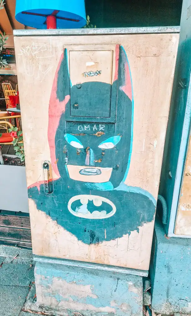 A weathered utility box painted with a colorful, stylized Batman face and chest emblem, with the word "OMAR" scratched into the forehead. A quirky find during 48 hours in Tirana’s creative corners.