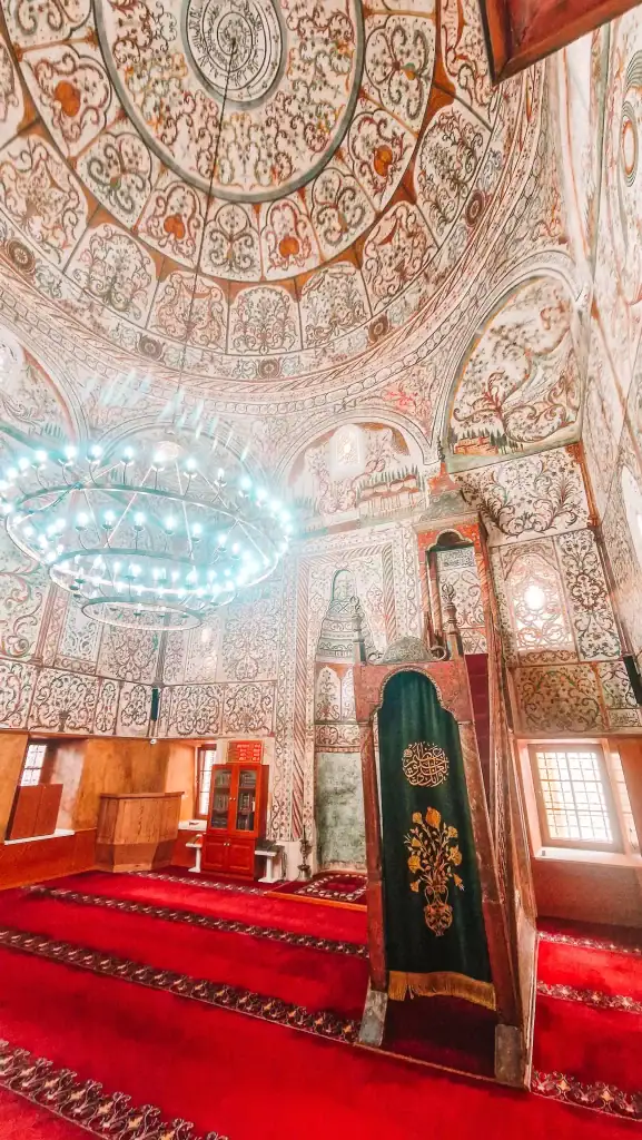 A richly detailed view inside the Et’hem Bey Mosque, with intricate floral and geometric ceiling frescoes, a grand chandelier, and a velvet-covered minbar standing on deep red carpet—one of the most beautifully preserved interiors you’ll see during 48 hours in Tirana.