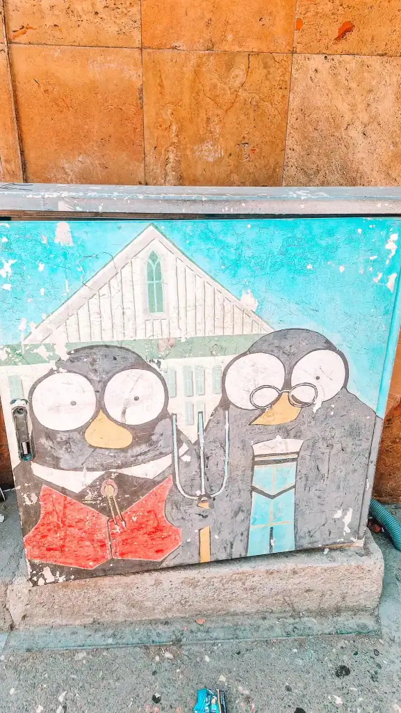 Whimsical street art on a utility box in Tirana featuring two cartoon penguins mimicking Grant Wood’s "American Gothic," complete with a farmhouse background and pitchfork—another fun visual you’ll stumble across during 48 hours in Tirana.