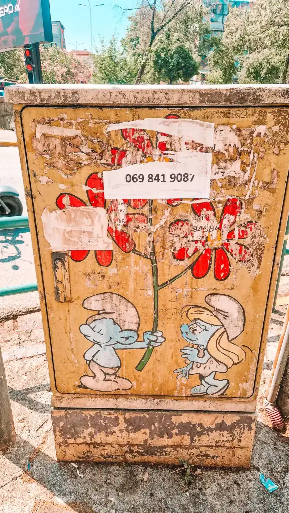 Faded mural of two Smurfs holding a giant red flower on a weathered yellow utility box, surrounded by peeled posters and graffiti—playful cartoon nostalgia right in the heart of Tirana’s streets.