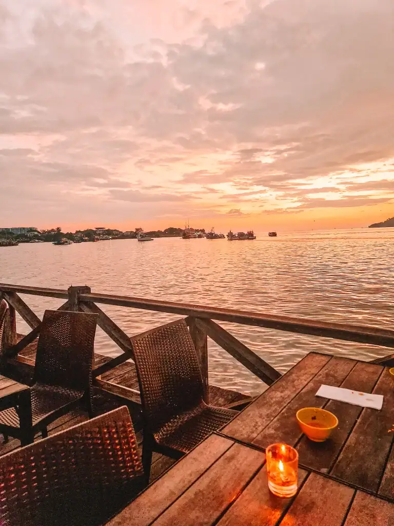 Warm candlelight glows on a wooden table at a seaside restaurant, with views of fishing boats and a fiery sunset over calm waters—perfect for winding down a day of exploring Malaysia.