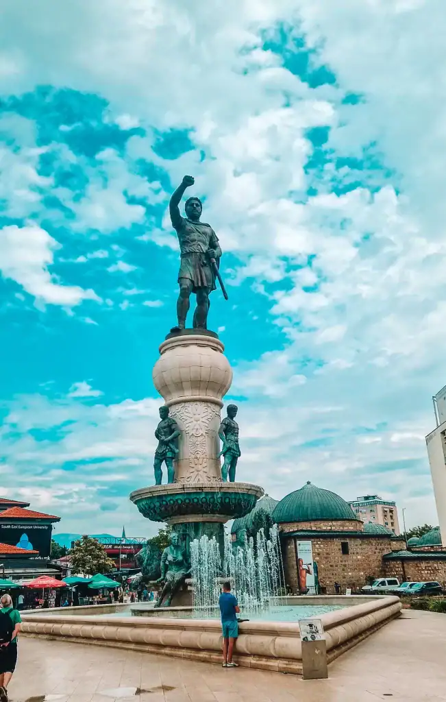 A towering statue of a warrior raising a fist stands atop a grand fountain in Skopje’s central square, with detailed carvings and multiple levels of sculptures surrounded by spouting water.