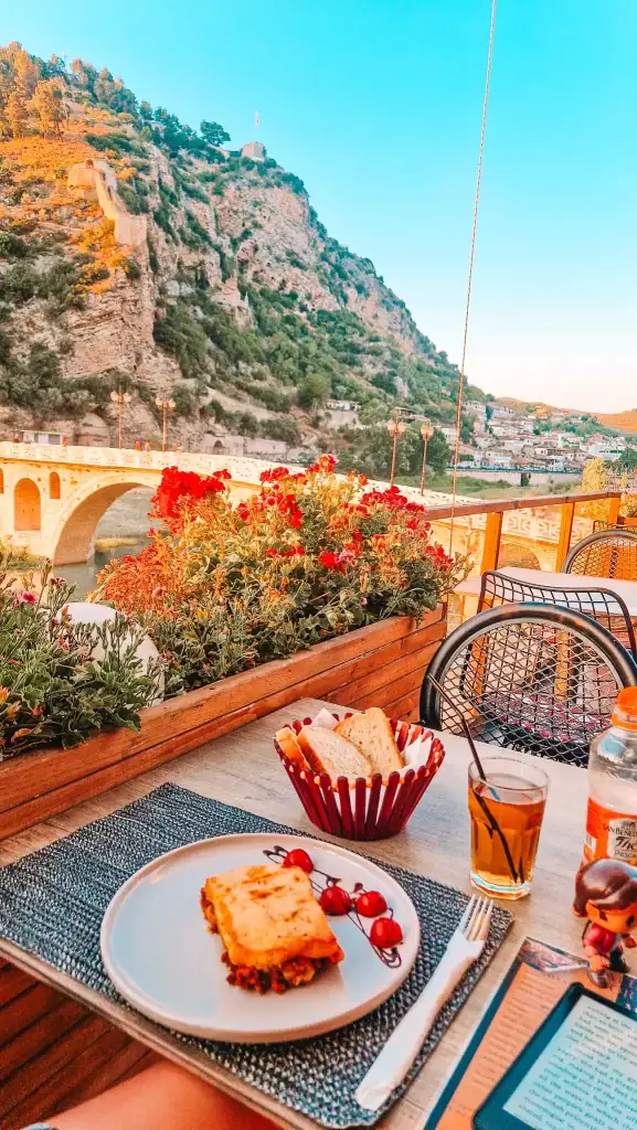Plate of moussaka with cherry tomatoes, breadbasket, and iced tea on a table overlooking a stone bridge, red flowers, and a mountain at sunset in Berat.