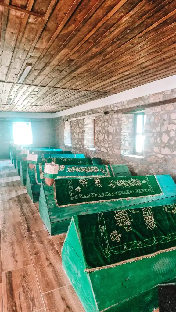 A row of green-covered tombs inside a stone-walled building with wooden floors and ceiling. Arabic script decorates the cloth on top of each tomb, illuminated by natural light from small windows.