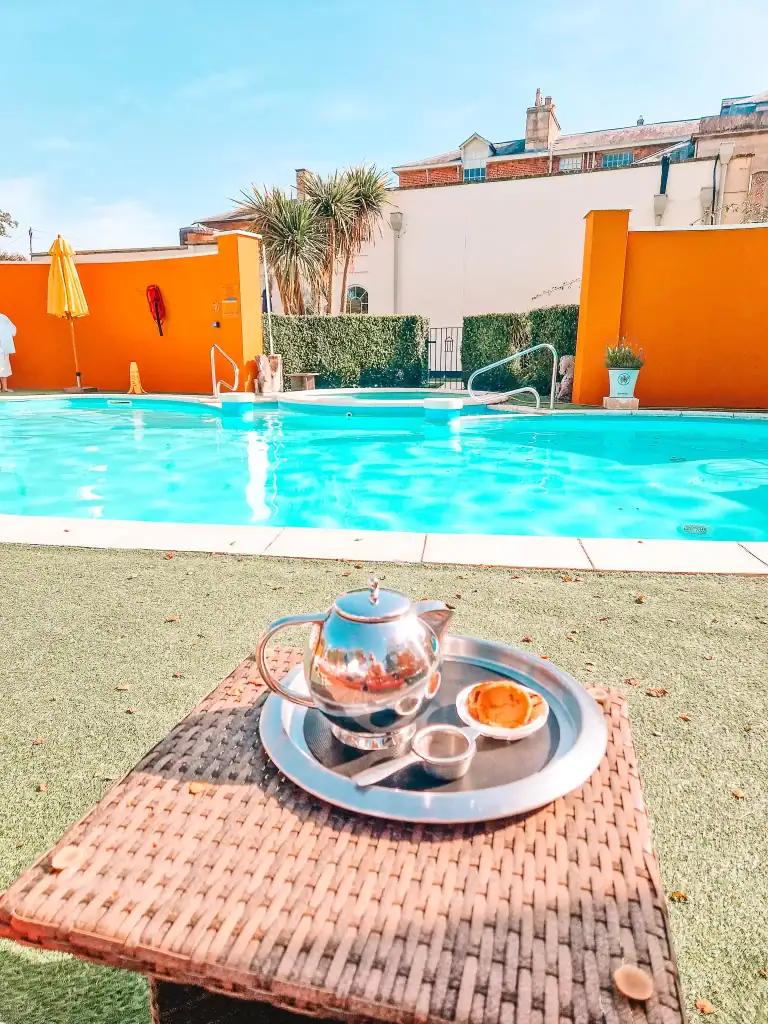 A silver teapot and a small tart sit on a tray atop a wicker table, set beside a bright blue outdoor pool with vivid orange walls and palm trees in the background. The scene suggests a relaxing sunny day at Bisshopstrow Hotel, Wiltshire.