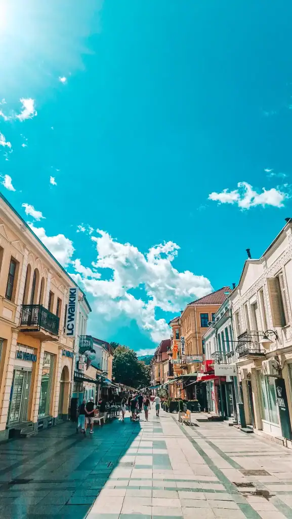 A sunny pedestrian street in Bitola, North Macedonia is lined with pastel-colored buildings and popular retail stores like LC Waikiki, with locals and tourists strolling.