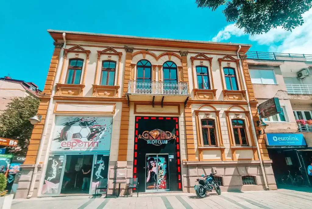 A historic two-story building in Bitola houses a slot club and betting shop, with ornate architecture and arched windows. A motorbike is parked out front.