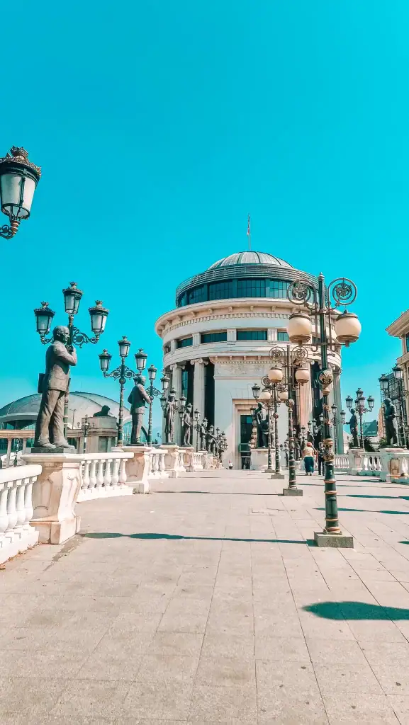 A neoclassical white building with a domed roof and grand columns stands at the end of a pedestrian bridge lined with ornate lamp posts and statues of historic figures.
