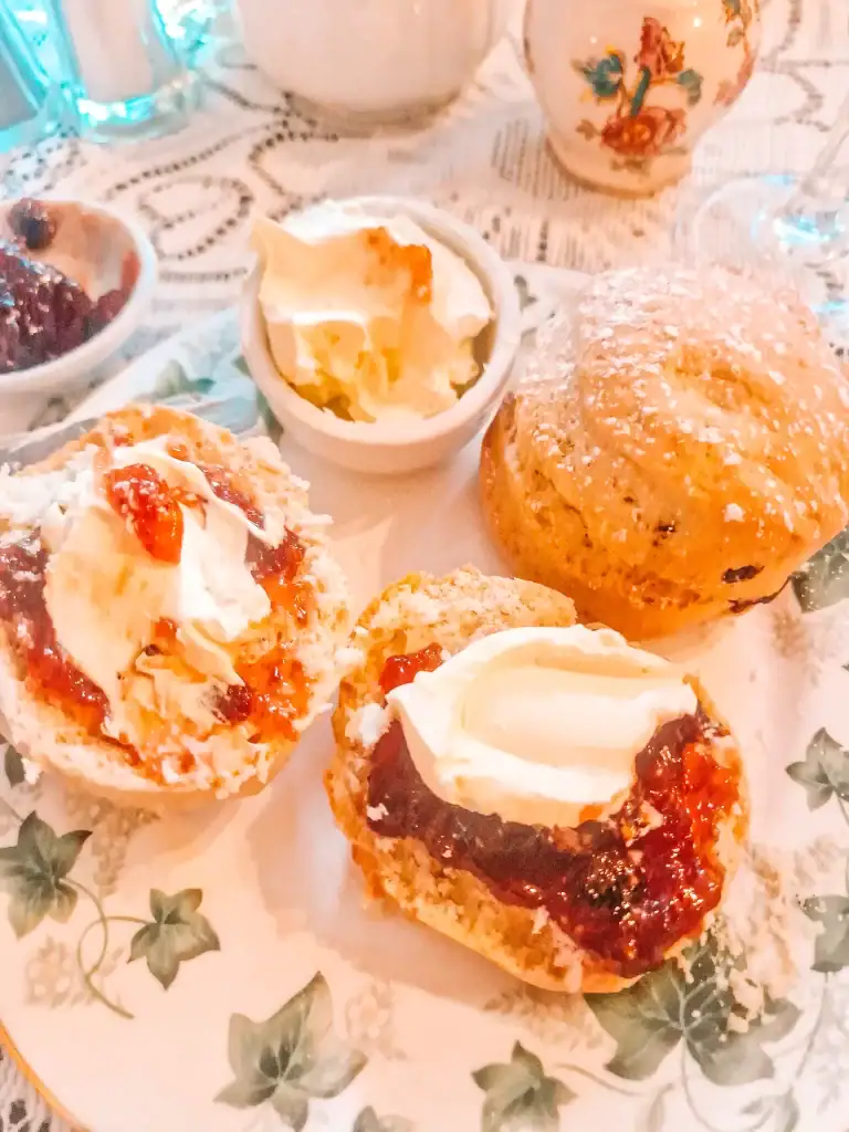 plate of scones topped with clotted cream and strawberry jam is surrounded by small dishes of cream and jam, all placed on a floral china plate with lace tablecloth underneath. A classic English cream tea setup.
