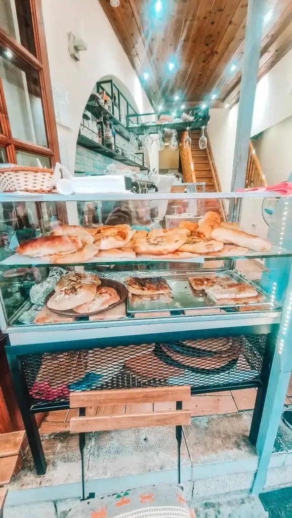 A glass display case inside a local bakery café in Gjirokaster, filled with savory pastries including byrek and flatbreads. Warm lighting and a wooden ceiling create a cozy indoor atmosphere.