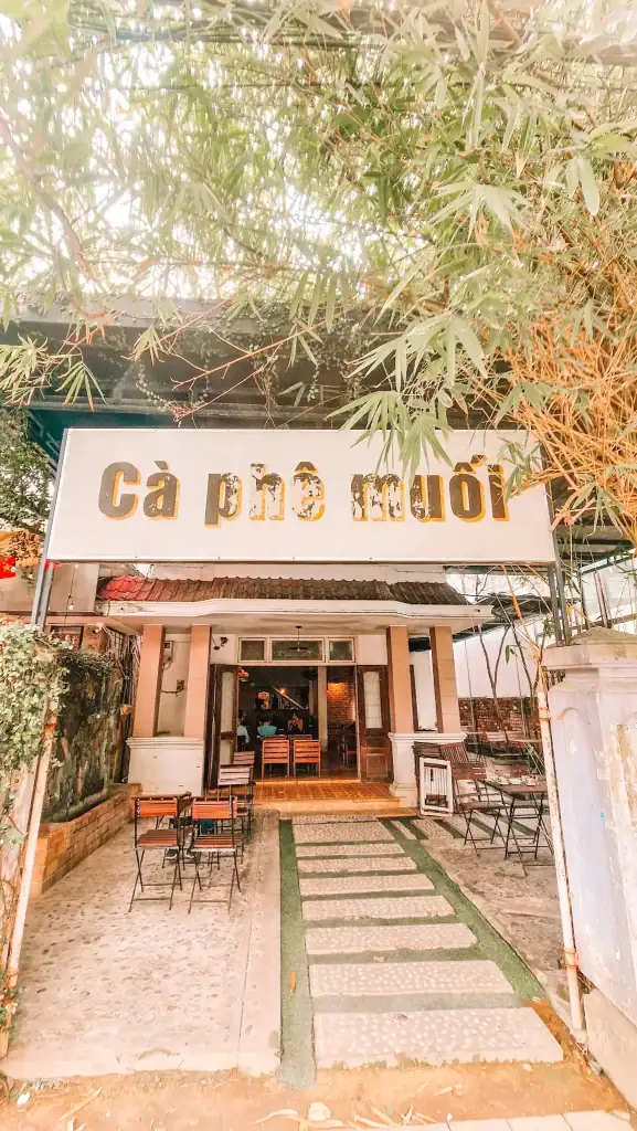 The front view of Cà phê muối, a cozy Vietnamese coffee shop with outdoor seating and a leafy canopy above. The faded yellow sign contrasts with the soft lighting and welcoming atmosphere.